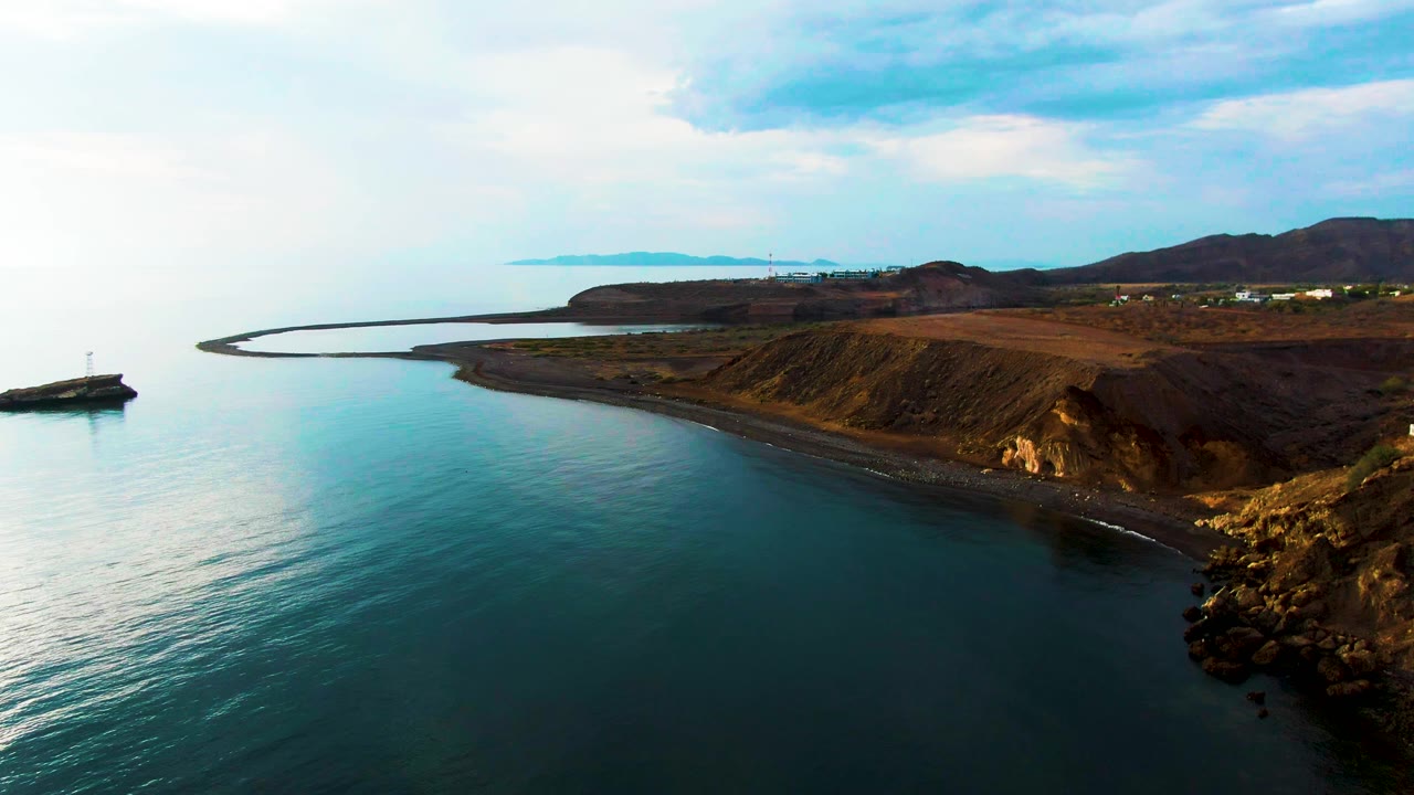Santa Rosalia coastline from above, with smooth blue water along a narrow dark beach, brown cliff faces on the right, distant hills, and no visible people.
