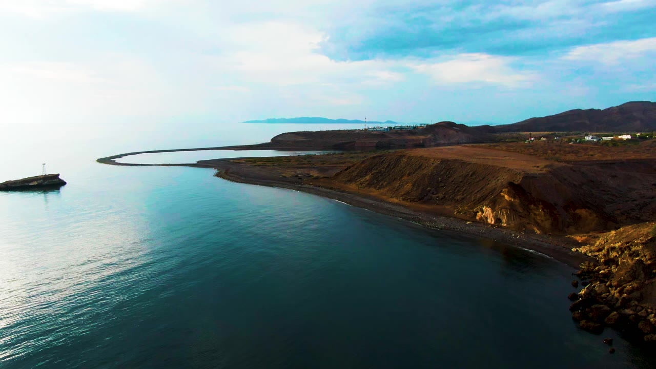 Aerial coastal view in Santa Rosalia showing still water in the foreground, a rocky cliff edge at right, the curved harbor wall in the background, and no visible people.