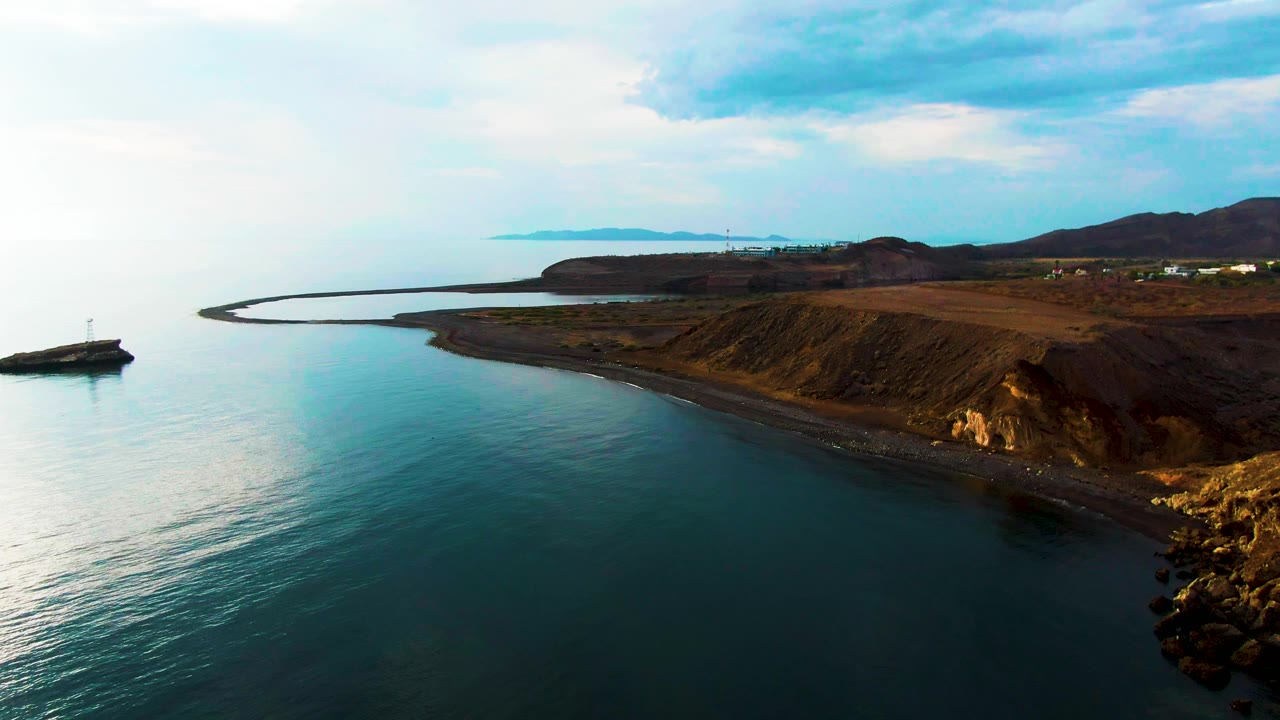 Wide aerial coastline with calm water and dry hills under cloudy sky.