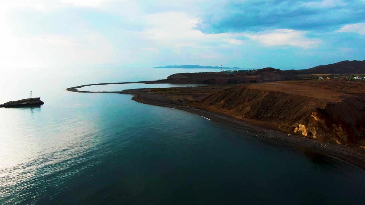 Aerial view in Santa Rosalia, Baja California Sur, with a small lighthouse on a rocky outcrop at left, calm blue-green water below, brown coastal cliffs at right, distant land on the horizon, and no visible people.