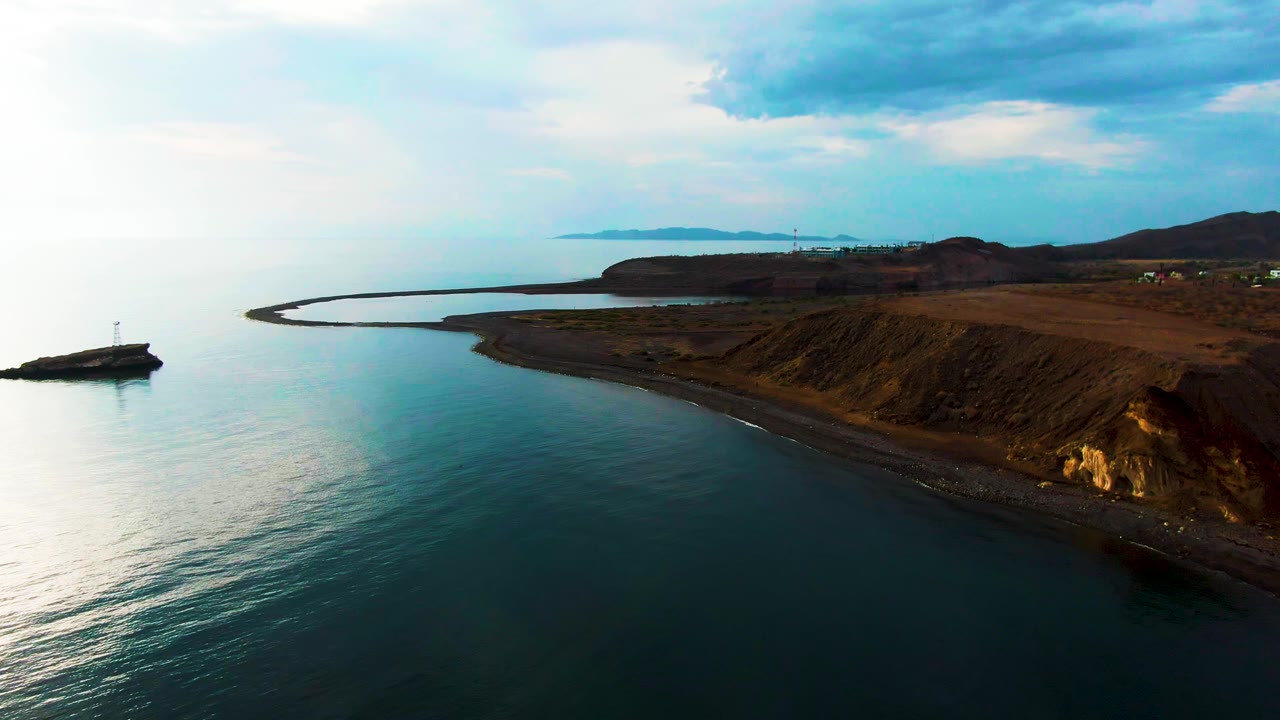 A wide aerial frame over Santa Rosalia coastline shows the lighthouse outcrop near the left edge, smooth ocean water in the foreground, a long curved shore, low brown hills, scattered clouds, and no visible people.