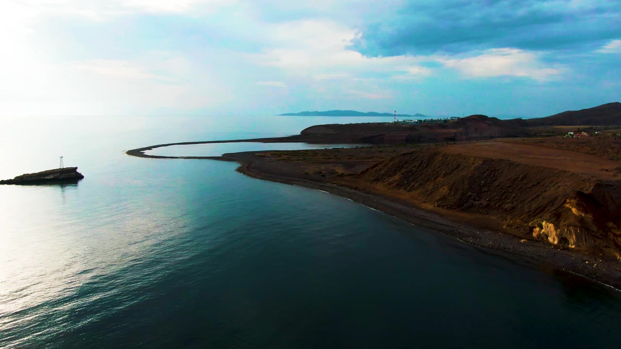 Santa Rosalia shoreline runs diagonally across the frame with calm water in front, the small lighthouse on its separate rock at left, darker clouds overhead, distant land beyond the bay, and no visible people.