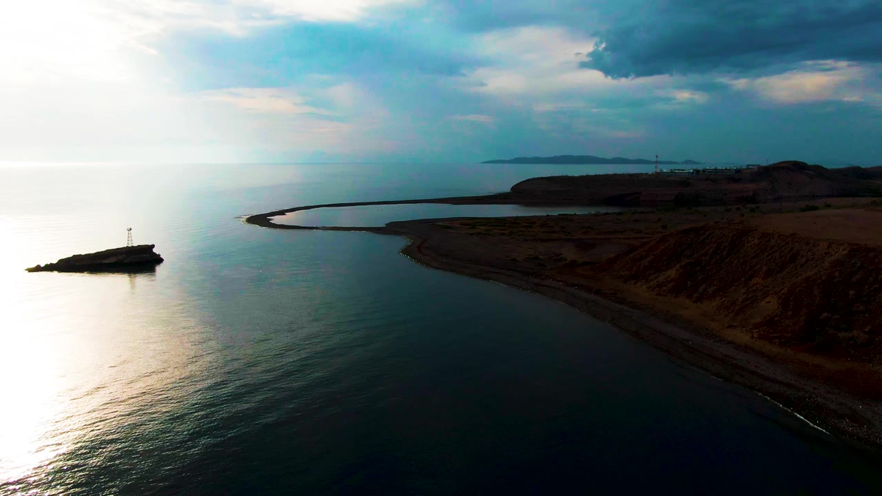 Santa Rosalia coast viewed from above, with the lighthouse outcrop near the left edge, bright reflected sunlight on the water, a dark cliff line to the right, distant land on the horizon, and no visible people.