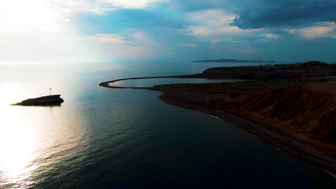 Santa Rosalia coastal view with the lighthouse outcrop low on the left, a vertical band of sunlight across the ocean, the curved beach and cliff shelf stretching right, layered clouds, and no visible people.