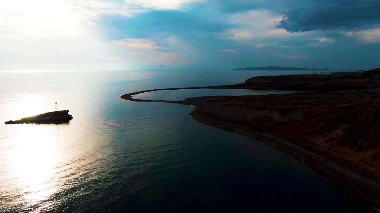 Aerial frame over Santa Rosalia, Baja California Sur showing a small rocky island with a lighthouse marker at left, a curved breakwater and dark shoreline at right, bright sun reflection on calm water, and no visible people.