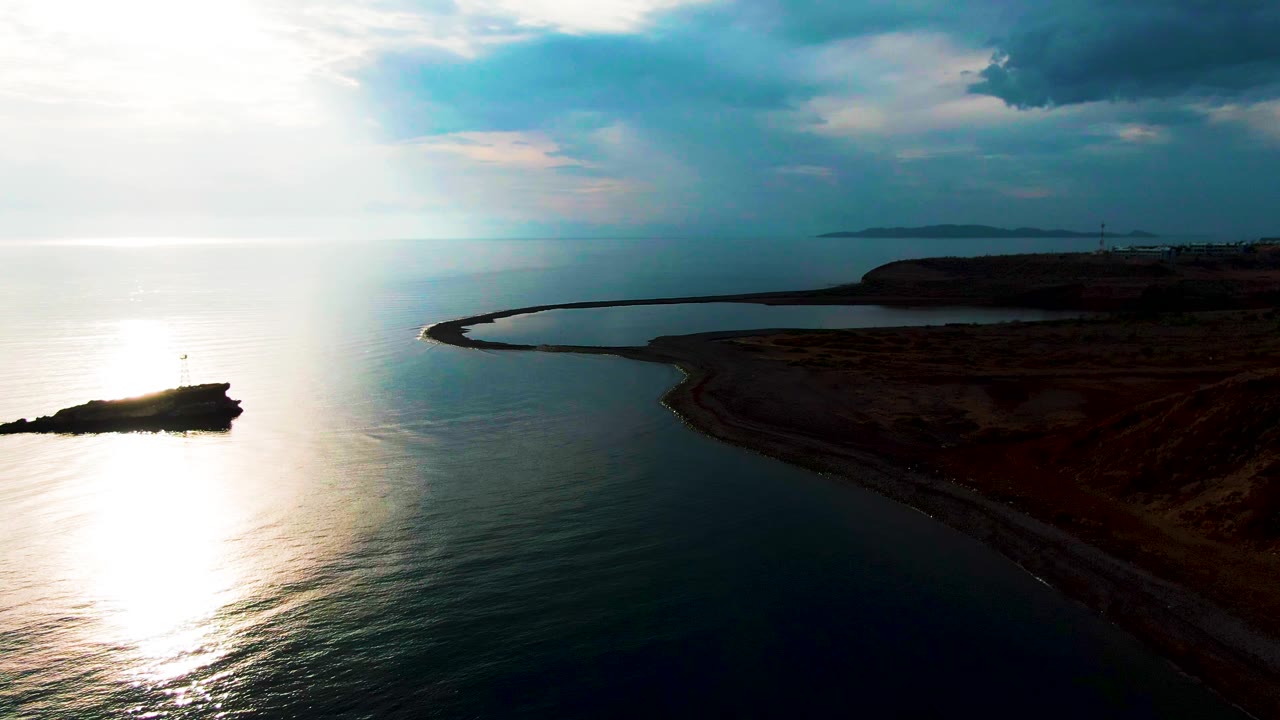 Wide aerial over Santa Rosalia with the rocky island at lower left, a smooth band of reflected sunlight on the water, the breakwater enclosing a darker inlet, heavy clouds above, and no visible people.