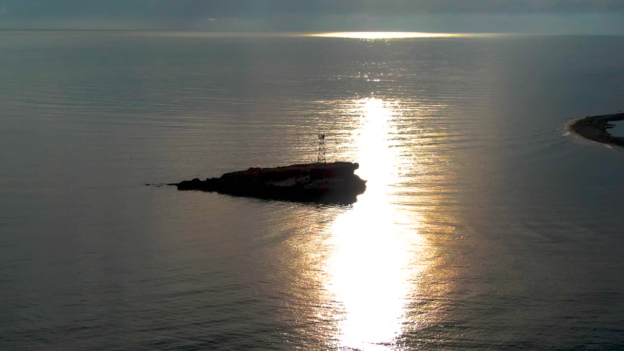 A rocky islet with a small lighthouse sits left of a bright vertical sun reflection on calm ocean water near Santa Rosalia, Baja California Sur, with no visible people.