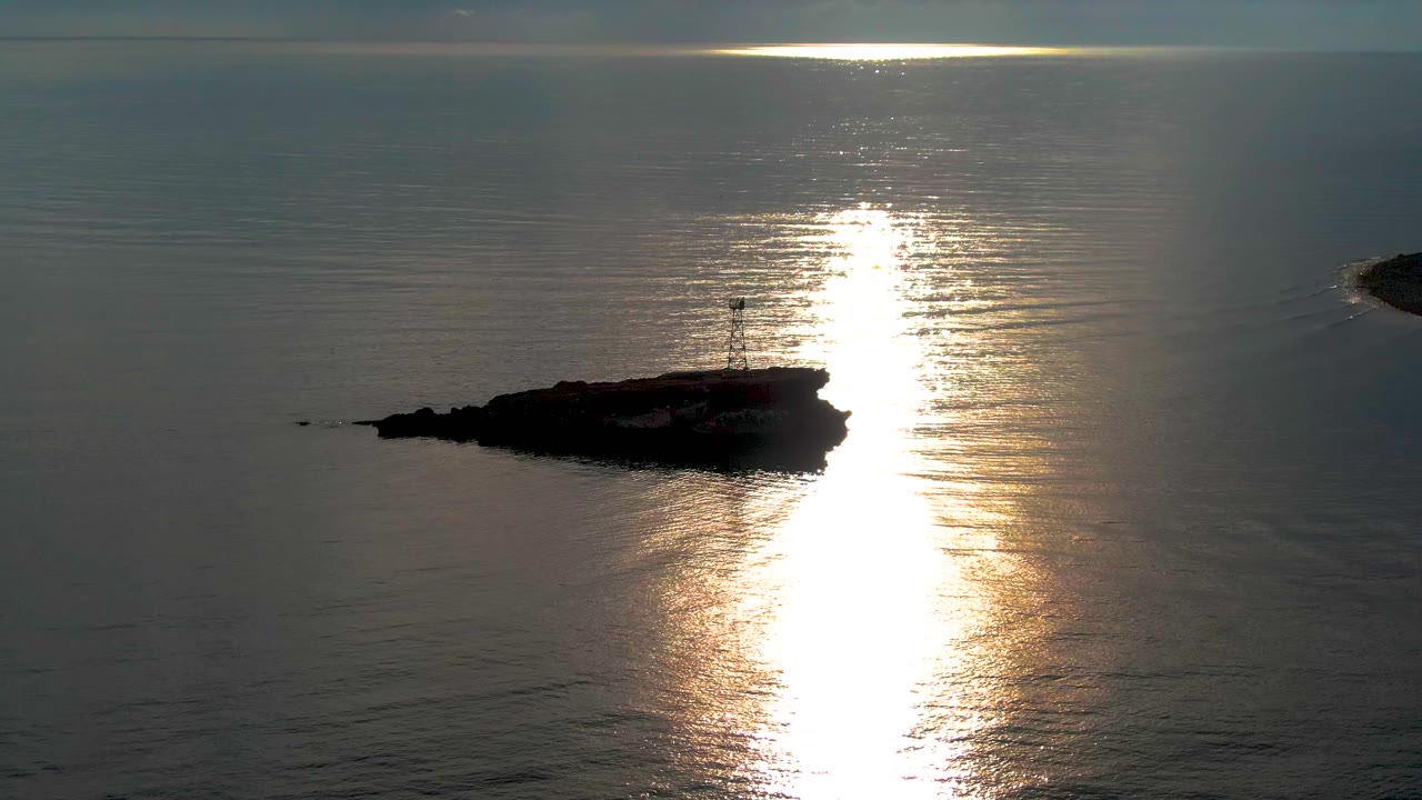 Small lighthouse on a dark islet beside bright reflected sun.