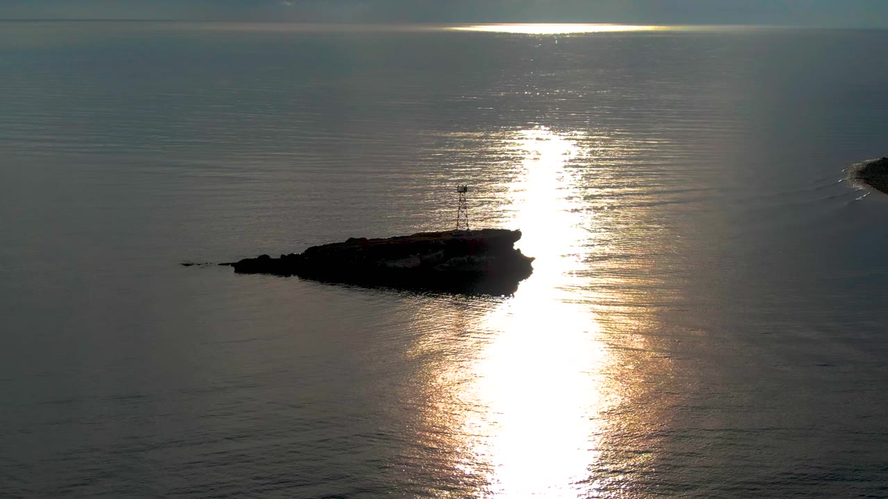 A low rocky islet with a small lighthouse appears against calm ocean near Santa Rosalia, while the sun reflection forms a broad shimmering strip across the water and no people are visible.
