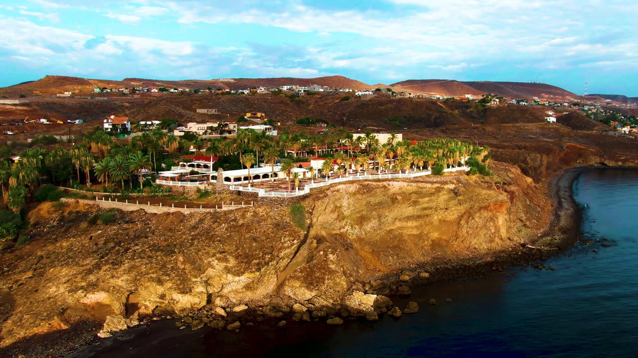 At 0:00, an aerial view in Santa Rosalia, Baja California Sur shows dark ocean along a rocky cliff, white cliff-top buildings, palms, brown hills, partly cloudy sky, and no visible people.