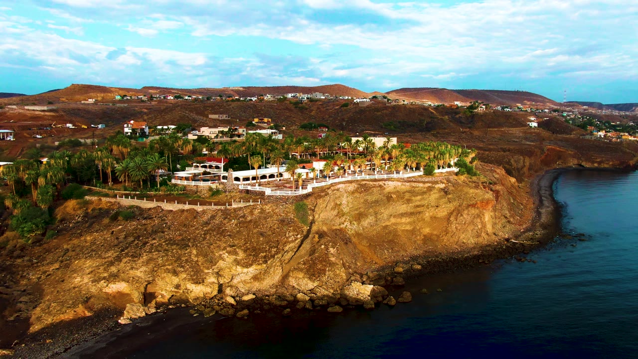 At 0:01, the Santa Rosalia coastline remains in a wide aerial frame, with the rocky waterline lower in view, the cliff-top property centered, dry hills behind it, and no visible people.