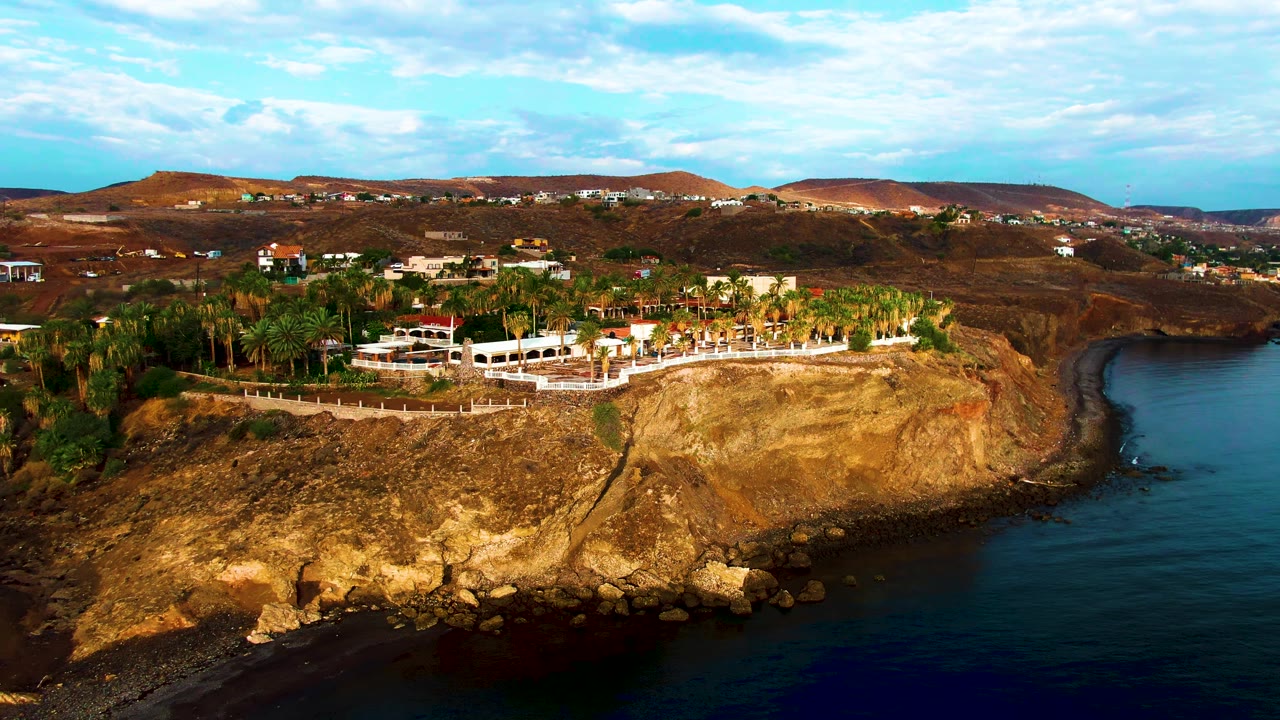 At 0:02, the aerial view shows more open water on the right side of the Santa Rosalia cliff, with tan rock, palms, white buildings, hillside homes, and no visible people.