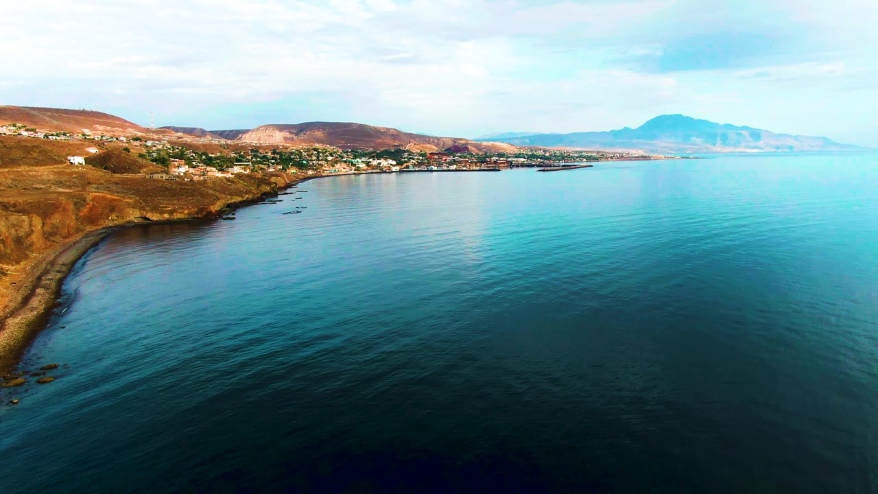 Opening frame of Santa Rosalia, Baja California Sur, Mexico, showing calm blue ocean in the foreground, a dry shoreline at left, scattered buildings, distant hills, and no visible people.