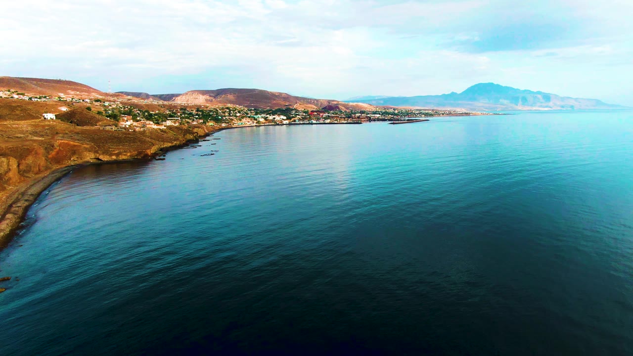 Calm ocean water curves along a dry hillside coast with small buildings and distant mountains.