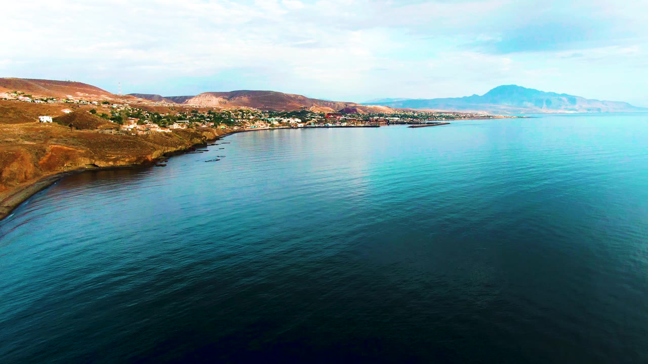 Aerial coastline view with turquoise water, low hills, scattered buildings, and distant landforms.