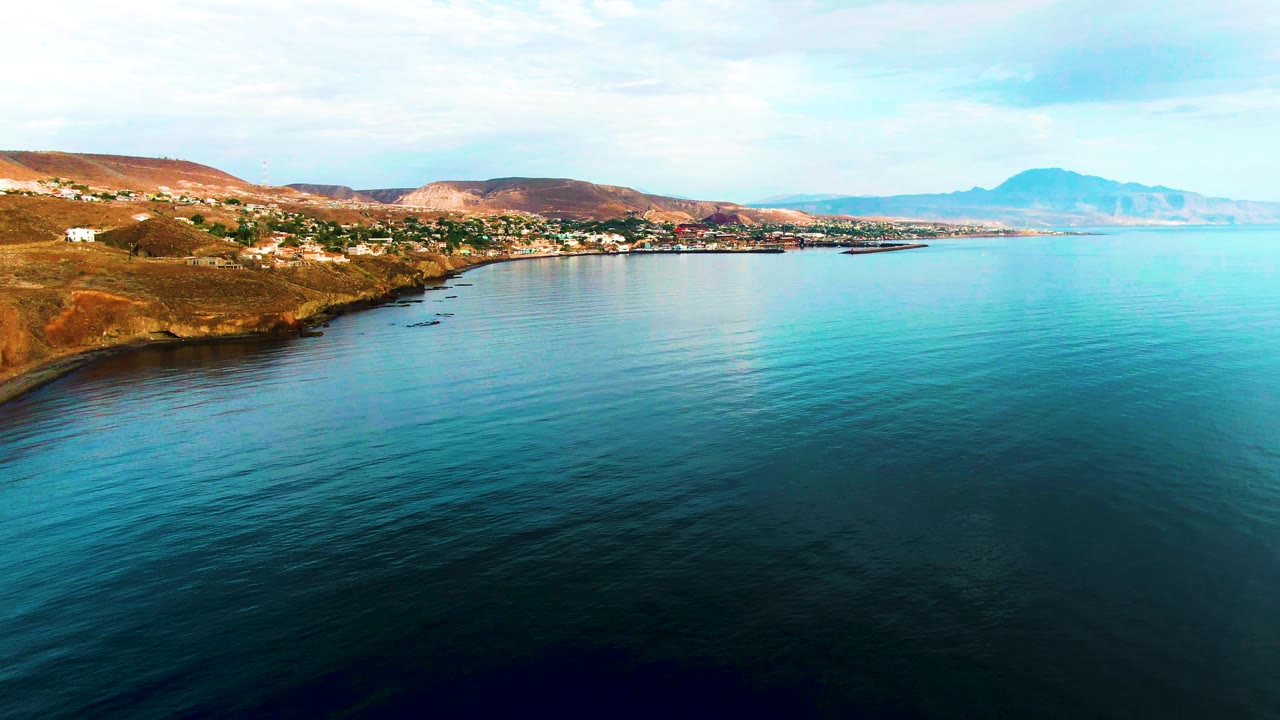 Opening aerial frame over Santa Rosalia, Baja California Sur, with calm blue-green water in front, the shore town across the middle distance, dry hills behind it, distant mountains on the right, and no visible people.