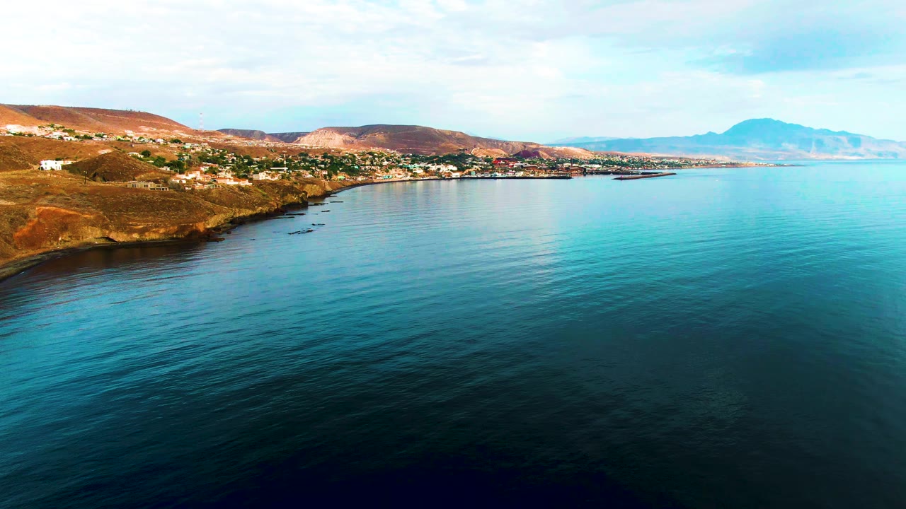 At 0:02, calm sea fills most of the foreground while Santa Rosalia's shoreline settlement sits beneath dry ocher hills, with a soft partly cloudy sky and distant mountain ridge visible to the right, and no visible people.