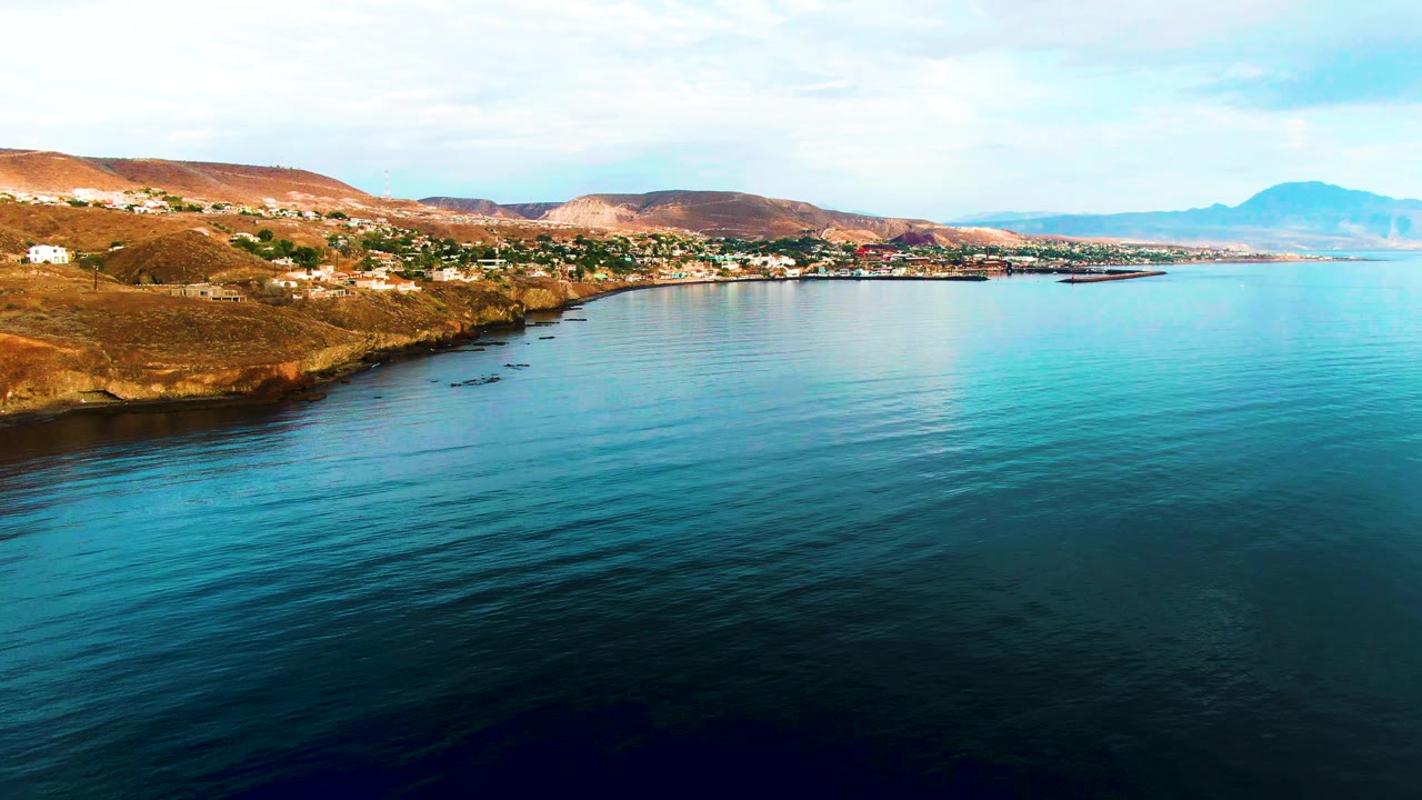 Wide aerial view of the Santa Rosalia, Baja California Sur shoreline, with calm blue ocean in the foreground, dry hills and scattered buildings along the coast, and no visible people.