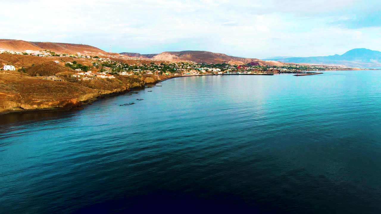 Santa Rosalia coastal panorama with dark blue water in the lower foreground, lighter turquoise water near shore, scattered white buildings on the left hillside, and no visible people.
