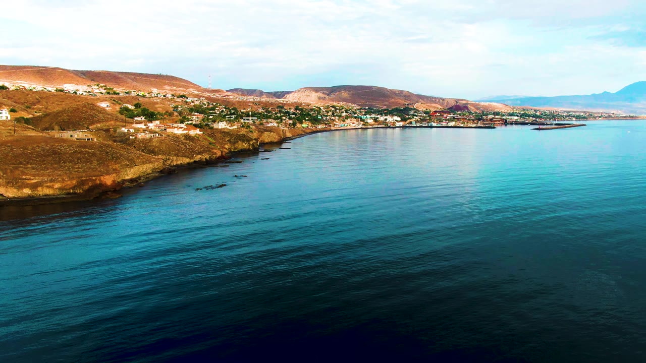 Wide aerial view over Santa Rosalia, Baja California Sur, with blue-green water in the foreground, rocky coast and hillside buildings beyond, and no visible people.
