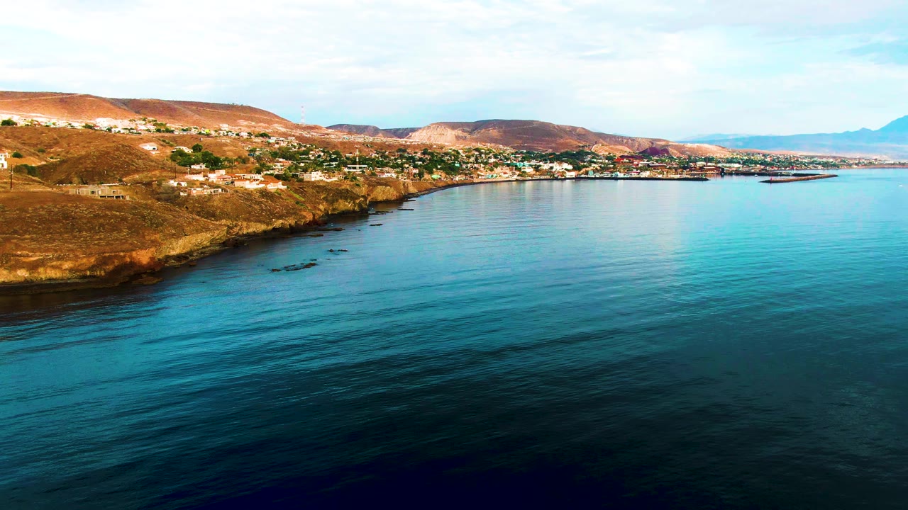 Aerial frame of Santa Rosalia coast with calm turquoise water stretching across the foreground, brown rocky shore at left, hillside buildings, and no visible people.