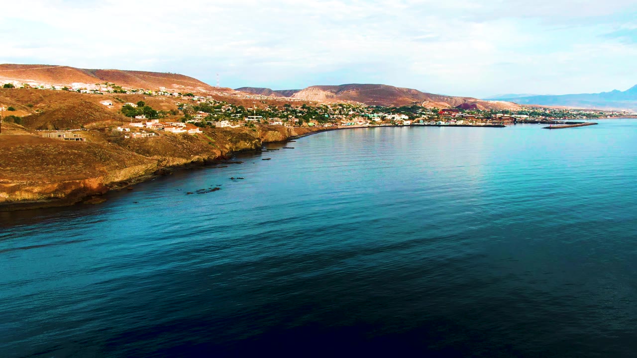 Santa Rosalia coastline seen from above, with a rocky point at left, small buildings along the hillside, broad blue water in front, and no visible people.