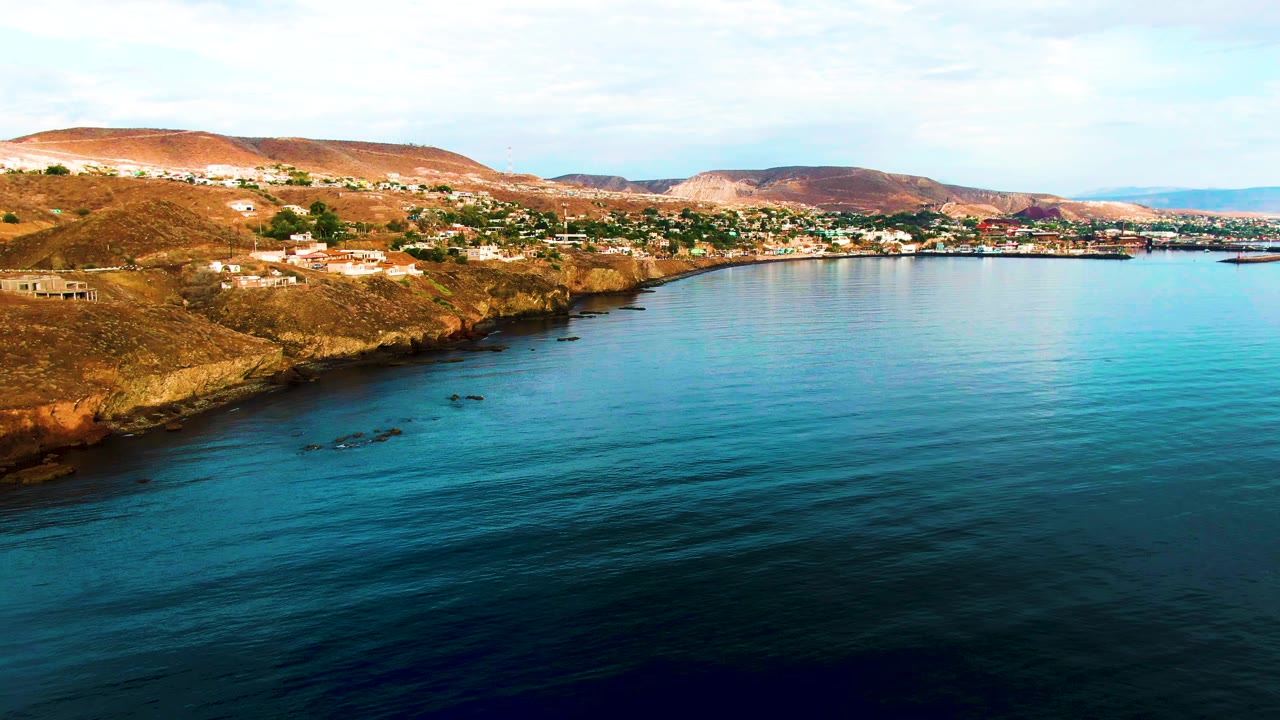 Aerial frame over Santa Rosalia, Baja California Sur, showing calm blue water, rocky brown shoreline, hillside buildings, distant hills, and no visible people.
