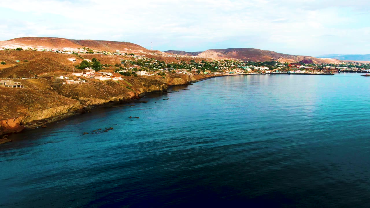 Second frame from the Santa Rosalia aerial view, with more open blue water across the foreground, dry shoreline at left, hillside buildings, distant rounded hills, and no visible people.