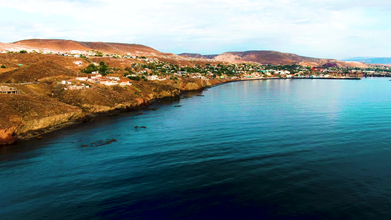 Santa Rosalia coastal aerial frame with a rocky brown shore running diagonally beside calm ocean, scattered hillside buildings, low hills in back, and no visible people.