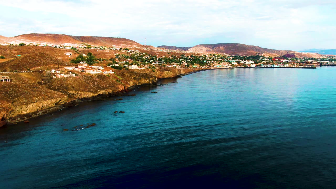 Aerial view of rocky shoreline, dry hillside town, and turquoise-blue water.