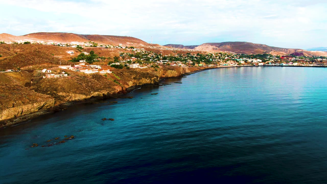 Opening aerial frame over Santa Rosalia, Baja California Sur, with calm blue ocean in the foreground, a rocky cliff edge, hillside houses, distant brown hills, and no visible people.