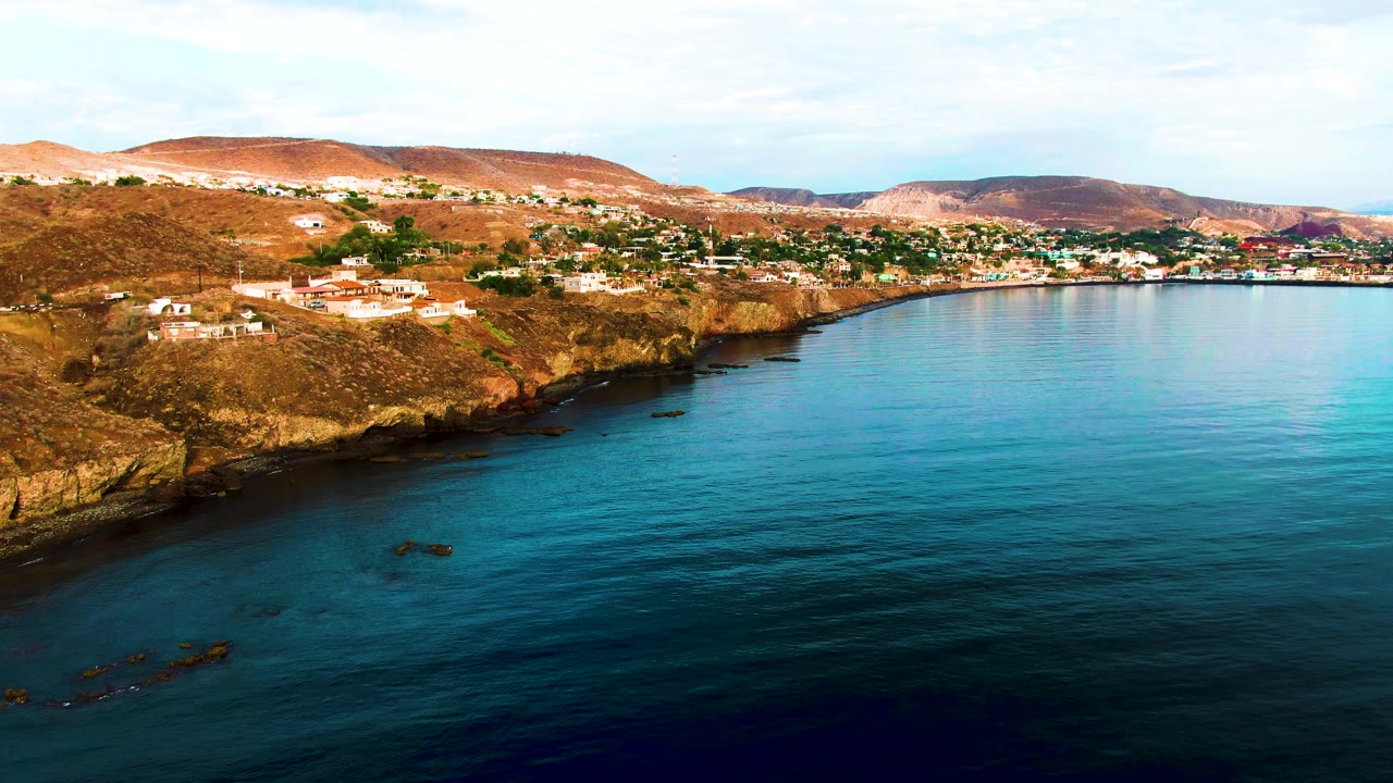 Santa Rosalia coastline seen from offshore, with darker blue water in the foreground, a rocky point on the left, hillside residences across the coast, distant hills, and no visible people.