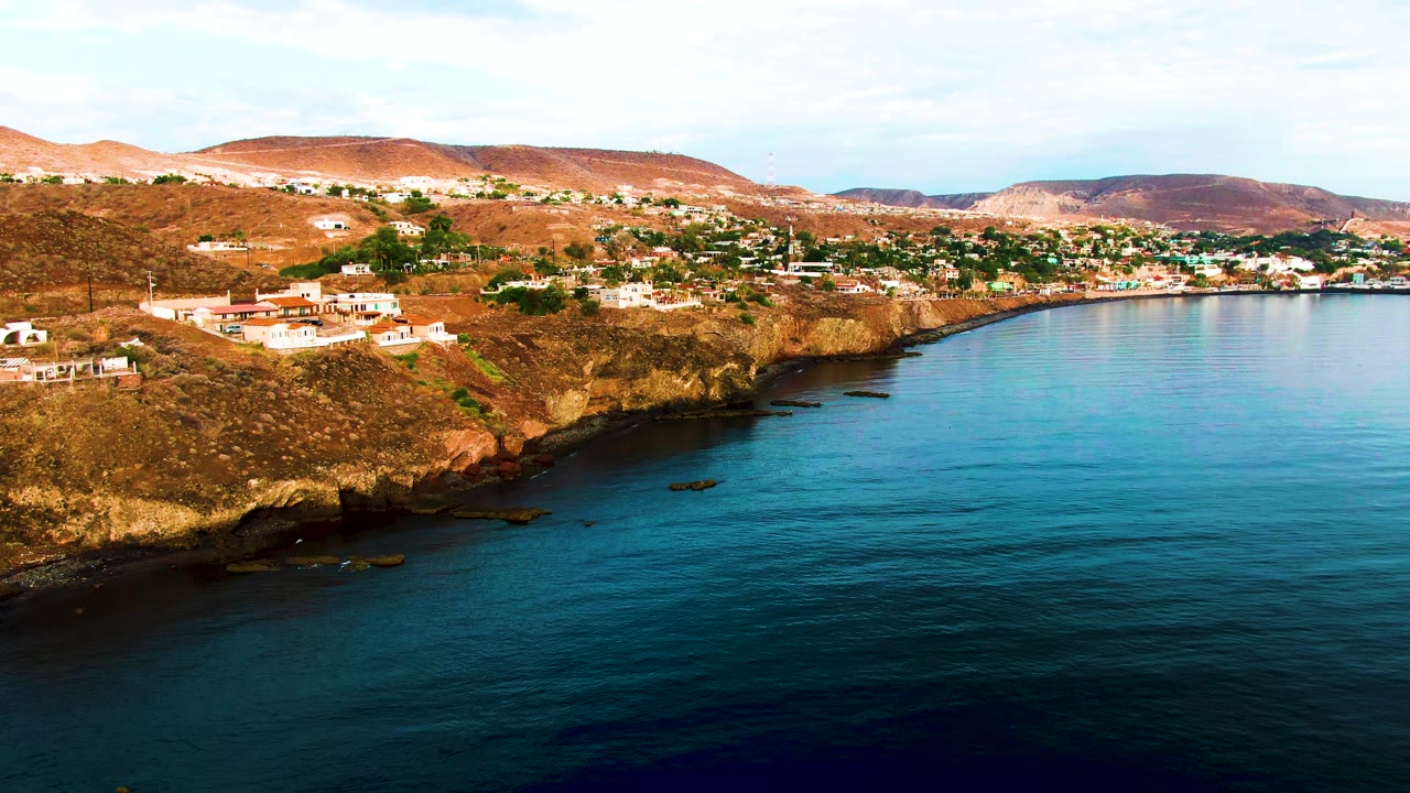 Opening aerial frame of Santa Rosalia, Baja California Sur, with white buildings on a brown coastal bluff, calm blue water below, dry hills behind, and no visible people.