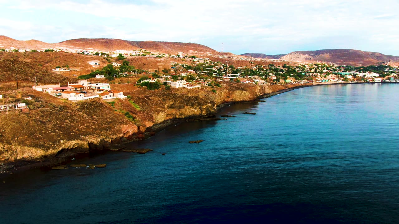 Santa Rosalia coastal aerial at 0:01, showing the cliff face stretching across the frame, white homes and red roofs on the hillside, blue ocean in front, and no visible people.