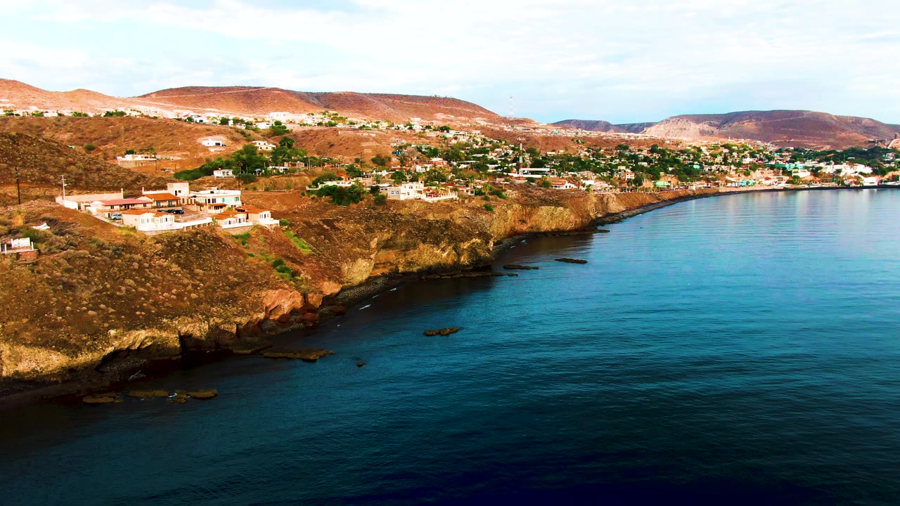 Santa Rosalia aerial frame with the coastline running diagonally, white buildings clustered along the bluff, turquoise-blue water at right and lower frame, dry hills behind, and no visible people.