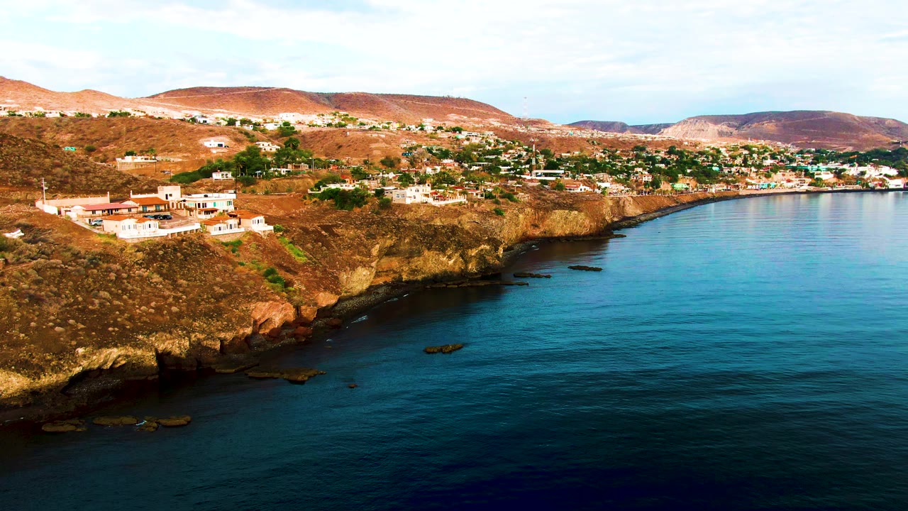 Aerial coastline view with white hillside buildings, rocky cliff shadows, and calm ocean.