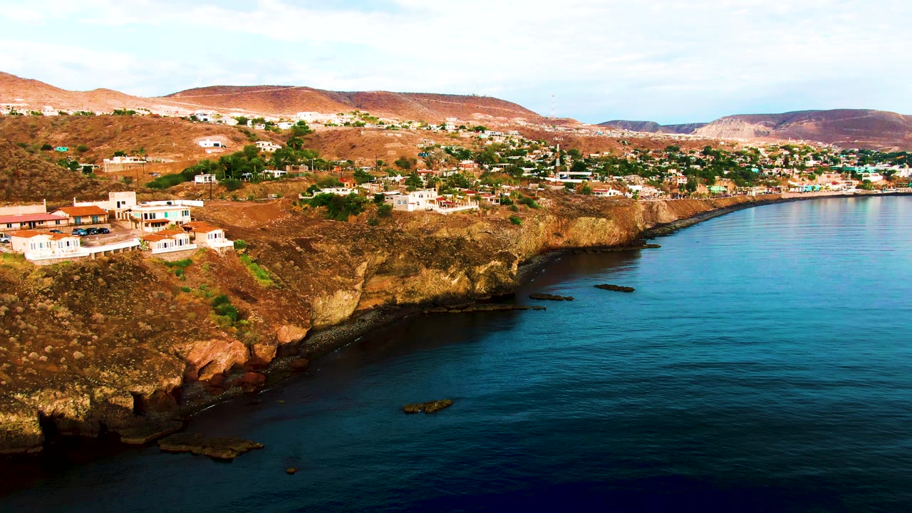 Aerial frame over Santa Rosalia, Baja California Sur, showing white buildings on a brown cliff above dark blue water, with dry hills behind and no visible people.