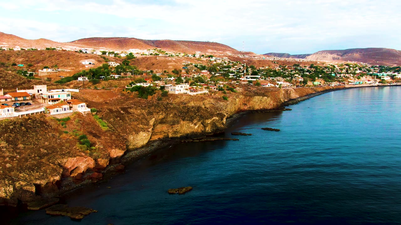 Aerial frame over Santa Rosalia with the left-side homes closer to frame edge, the curved rocky shore below, blue sea to the right, dry hills beyond, and no visible people.