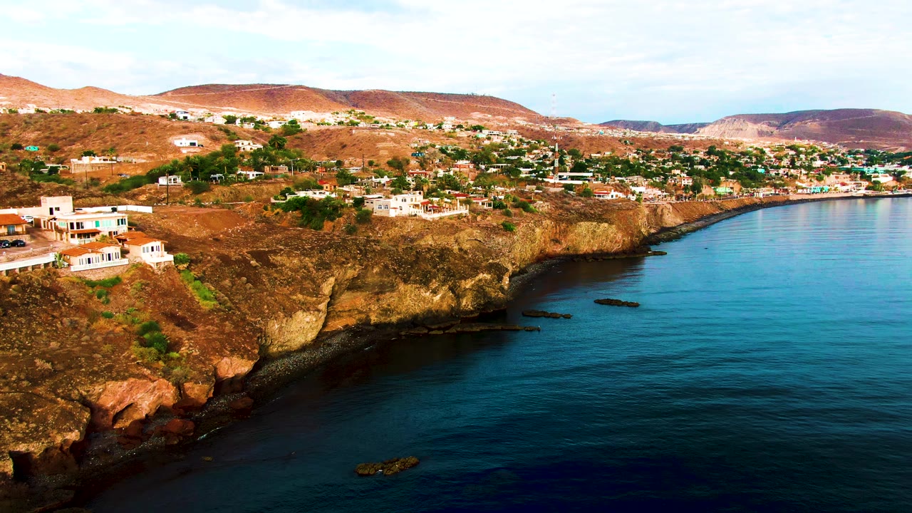 Aerial frame over Santa Rosalia showing more of the blue bay on the right, a line of cliffside homes across the upper frame, rocky brown slopes, and no visible people.