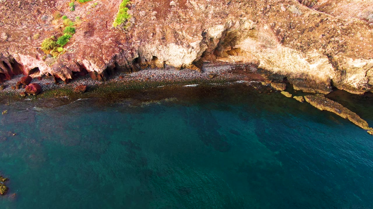 Aerial frame over Santa Rosalia coastline showing pale cliff rock, red shoreline stone, clear blue water, and a small light boat near the shore with no visible people.