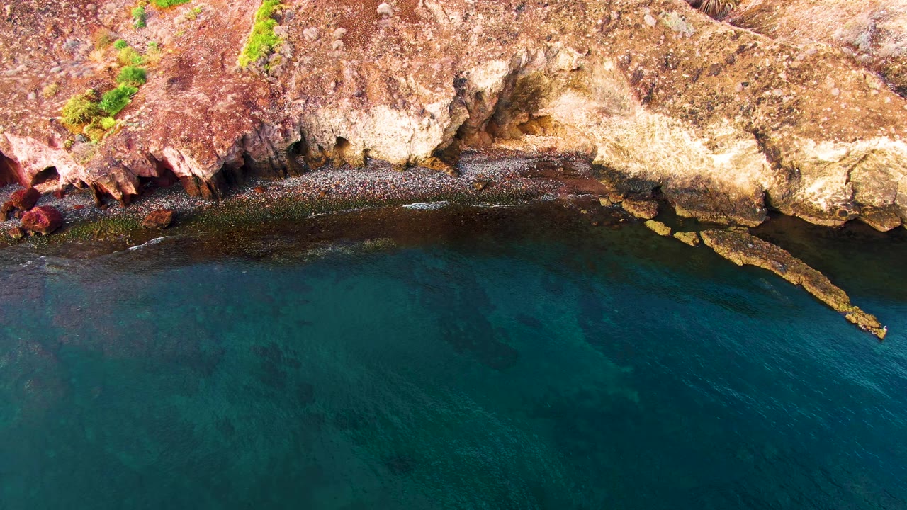Santa Rosalia rocky shoreline seen from above, with a narrow tan rock point extending into dark turquoise water and no visible people.