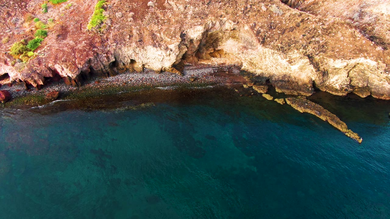 Aerial Santa Rosalia coastal frame with rough tan cliff faces, red rocks along the shore, dark shallow water near land, and no visible people.