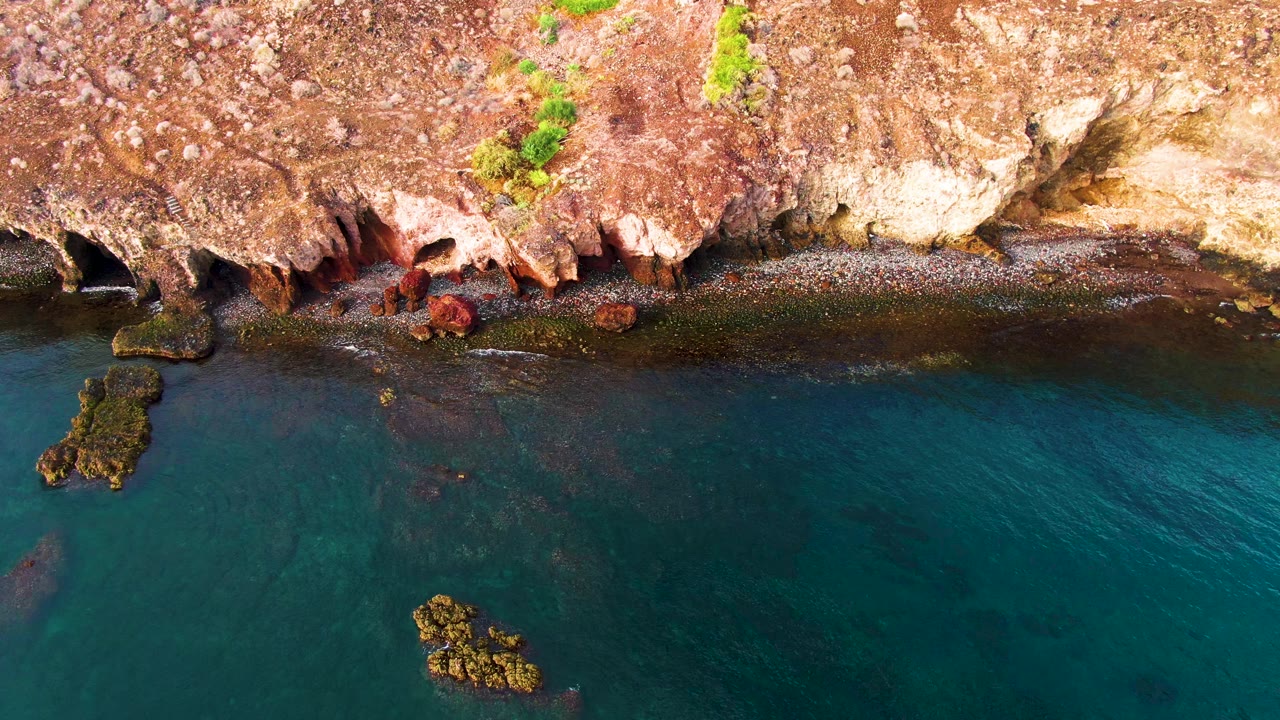 Aerial frame of Santa Rosalia coastline showing rust-colored rock, a dark shallow-water strip, blue-green sea, a small rock cluster near the lower center, and no visible people.
