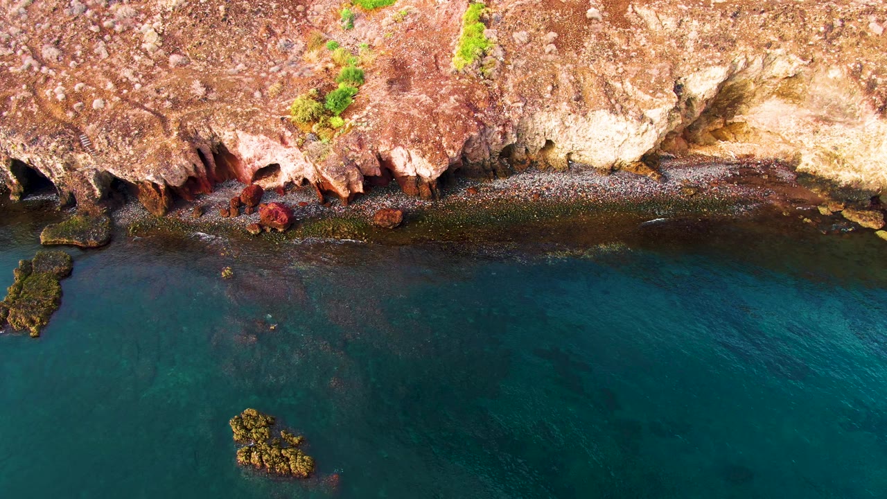 Top-down Santa Rosalia coastal view with pale cliff rock on the upper right, reddish shoreline rock on the left, transparent turquoise water, offshore stones, and no visible people.