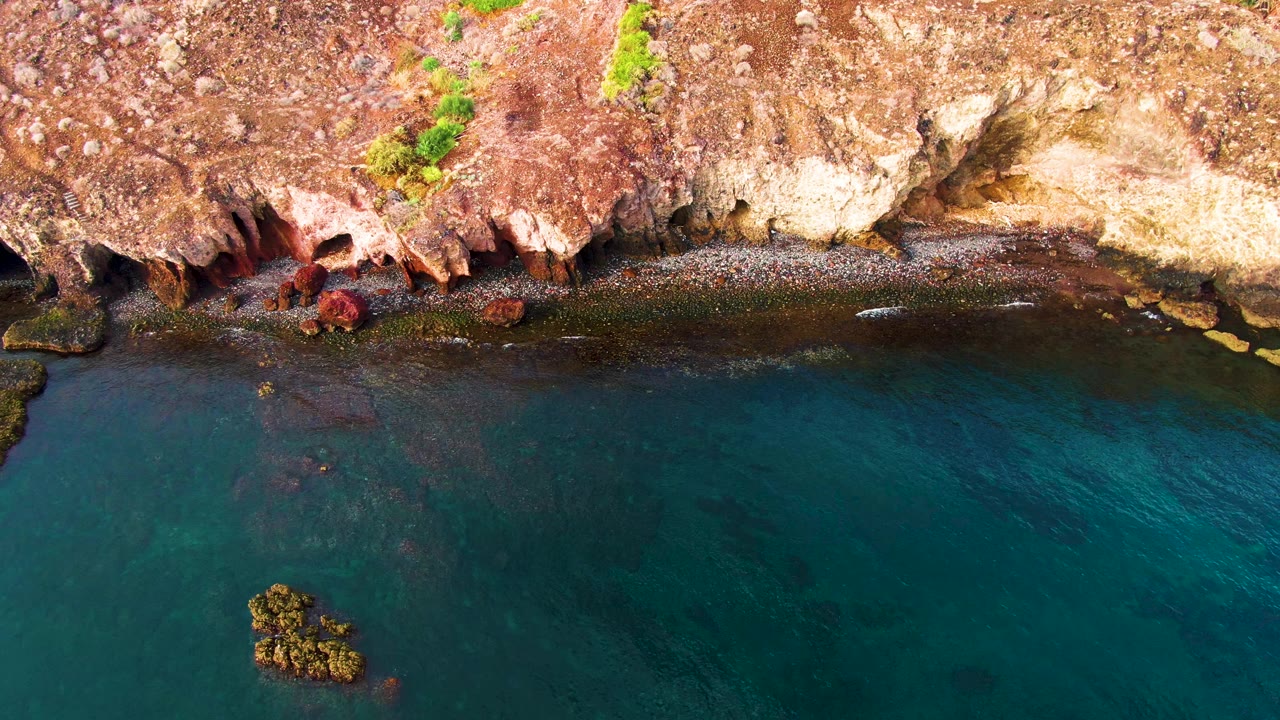 Overhead view of Santa Rosalia shore with a long dark waterline, small white wave marks, reddish cliff texture, a rock cluster in the lower water, and no visible people.