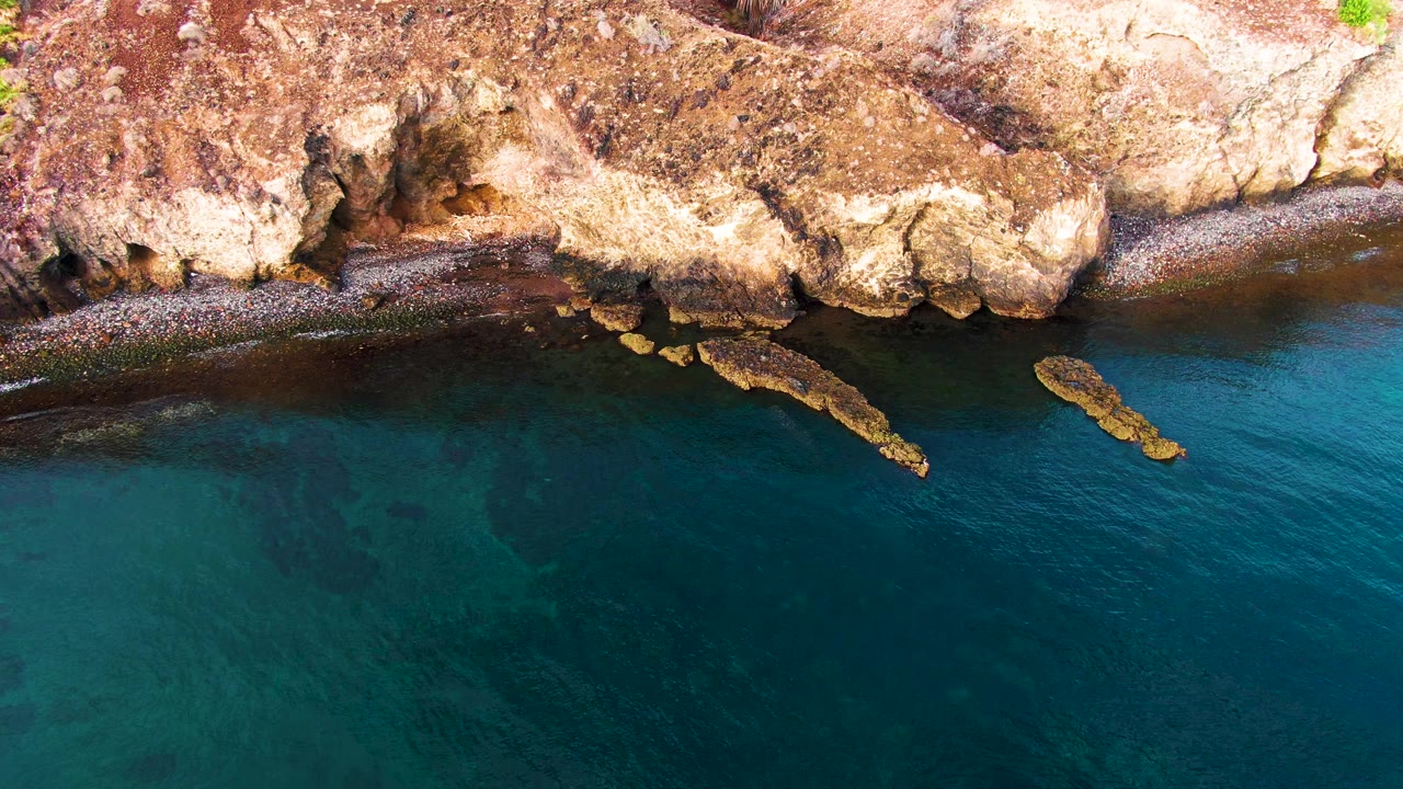 Aerial frame over Santa Rosalia coastal rocks and blue-green water, with the small bird slightly farther right near the shoreline and no visible people.