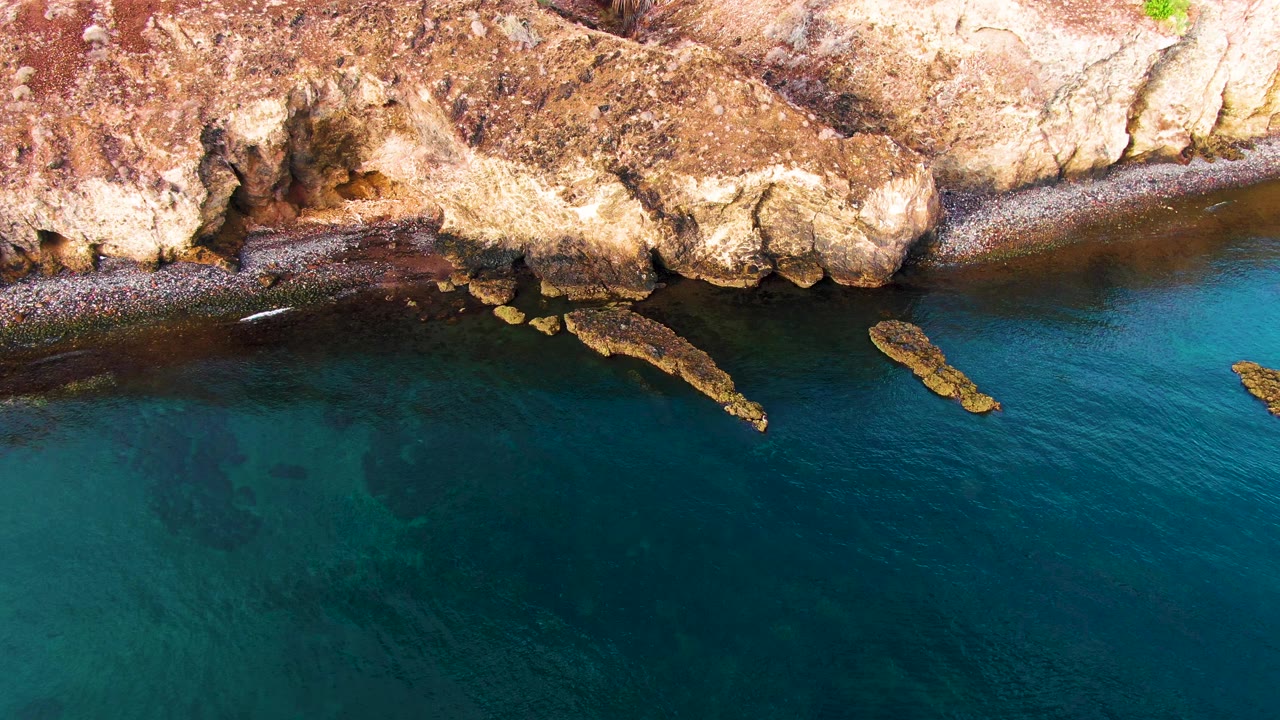 Aerial view in Santa Rosalia with rough tan rocks, a small bird low over rippled turquoise water, and no visible people along the shore.