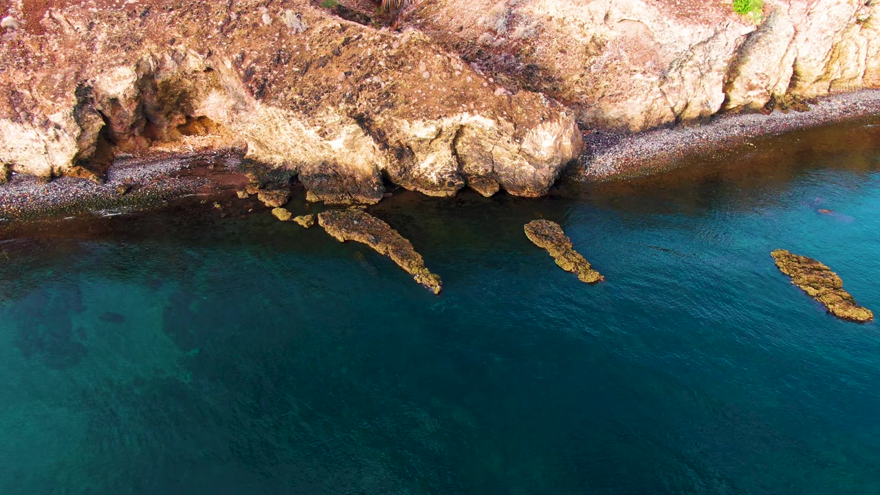 Overhead view of the Santa Rosalia rocky coast at the opening moment, with tan cliffs, a narrow pebbly shore, clear blue water, two offshore rock outcrops, and no visible people.