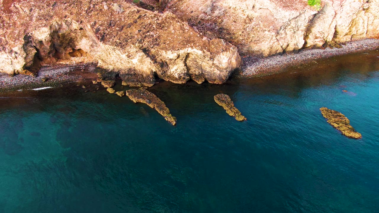 Aerial view of Santa Rosalia coastline one second in, with the darker water band near shore, bright tan rock ledges along the top, exposed offshore rocks, and no visible people.