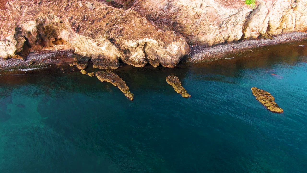 Overhead Santa Rosalia coastal frame with rust-colored cliff texture, a thin shore, turquoise patches in the water, a small bright offshore rock at right, and no visible people.